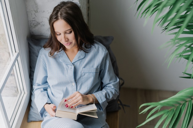 Woman with dark long hair, focused into book, reads something exciting, enjoys cozy atmosphere at home