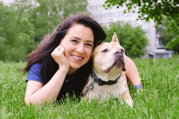 Woman with dark long hair is lying on the lawn with a dog smiling and looking at the camera