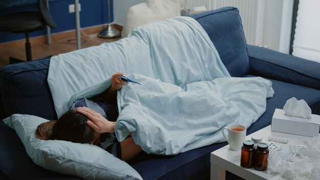 Woman with disease laying on couch and measuring fever using thermometer. Sick adult looking at device and checking temperature while having tissues and bottles of medicaments on table.