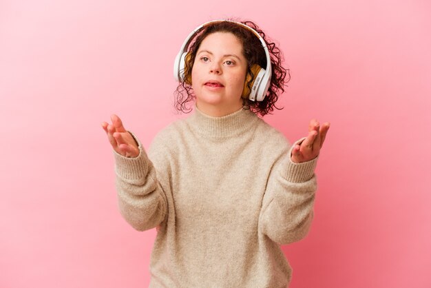 Woman with Down syndrome with headphones isolated on pink background receiving a pleasant surprise, excited and raising hands.