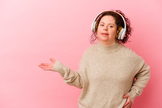 Woman with Down syndrome with headphones isolated on pink wall showing a copy space on a palm and holding another hand on waist