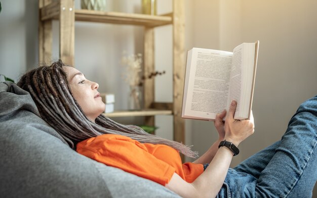 Photo woman with dreadlocks and bright clothes is sitting comfortably in a bag chair and reading a book