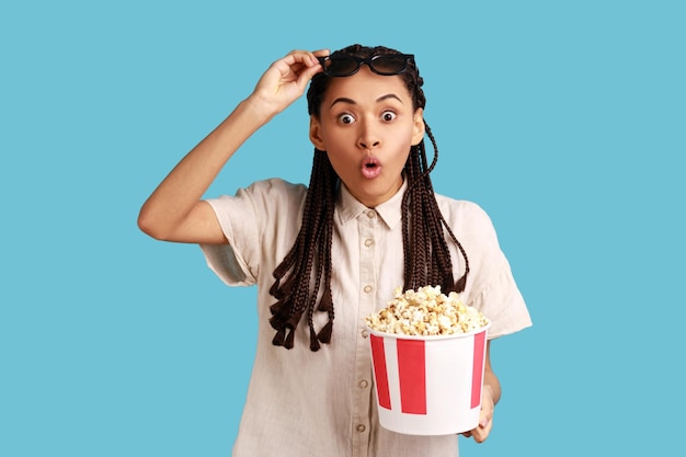 Woman with dreadlocks holding bucket popcorn and raising in 3d\
glasses sees shocked movie scene