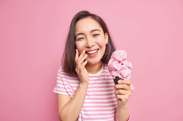 woman with eastern appearance smiles toothily dressed in casual striped t shirt holds waffle ice cream being in good mood 