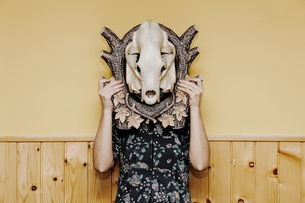 woman with face covered by animal skull standing against wall