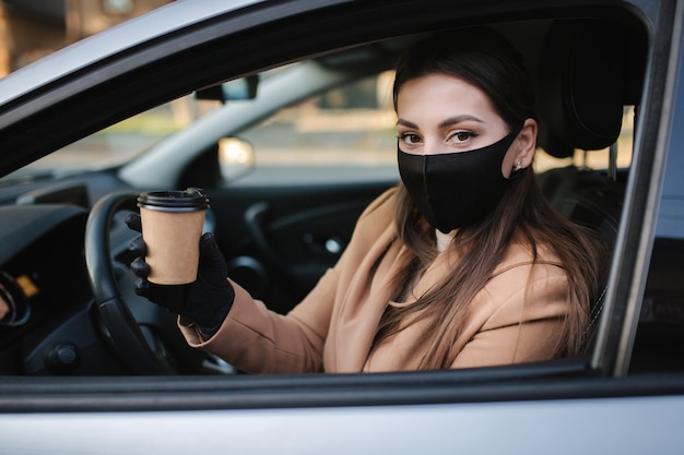 コロナウイルスのパンデミック中にコーヒーを飲みながら車を運転しているフェイスマスクの女性、