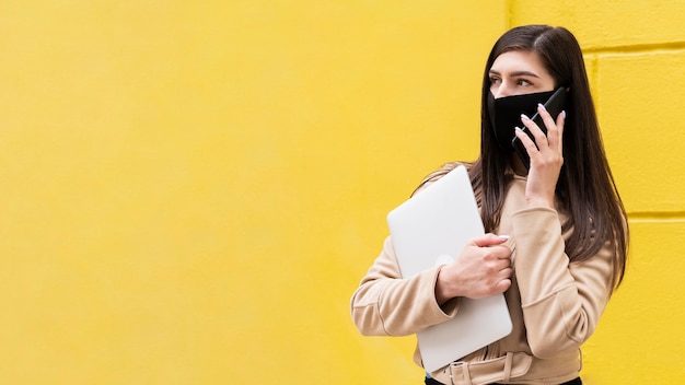 Photo woman with face mask holding laptop and talking on smartphone