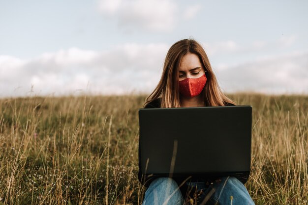 Woman with face mask working on laptop from remote location in natur