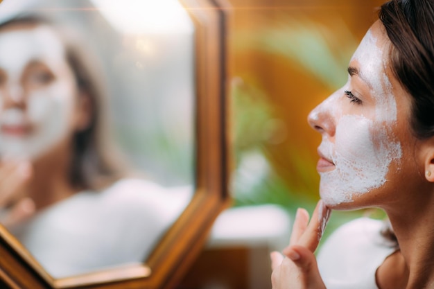 Donna con la maschera per la cura della pelle di bellezza facciale che guarda allo specchio a casa