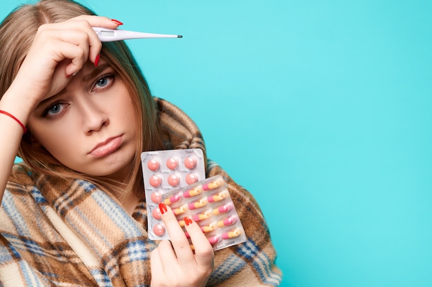 Foto la donna con la febbre e un termometro in mano beve pillole e si avvolge in un plaid