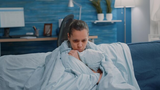 Woman with fever using blanket and pillow for warmth. Sick person shivering and having chills because of temperature and coronavirus symptoms, trying to get warm and cure sickness.