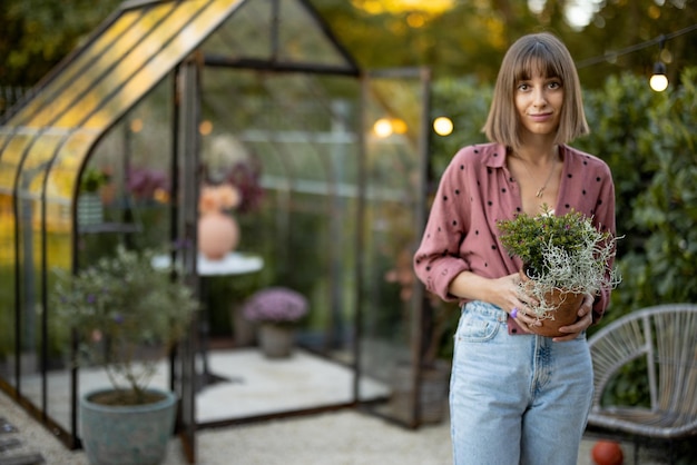 後ろに温室のある庭で植木鉢を持つ女性