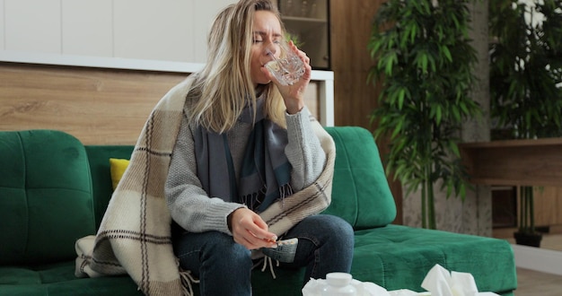 Woman with flu drinking medicine on sofa at home against cold illness disease sickness. Concept of healthcare and illness.