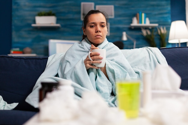 Фото Женщина с гриппом сидит дома, пьет горячий чай, чувствуя себя больным, человек с температурой принимает лекарство ...