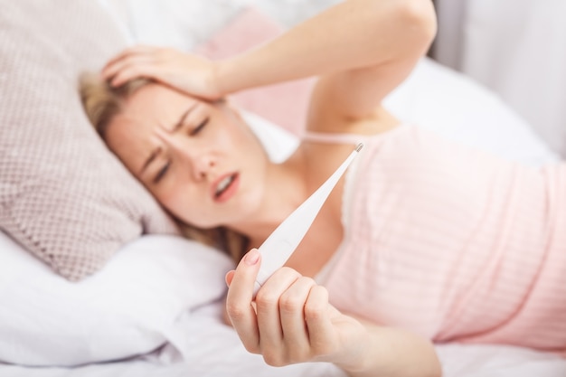 Woman with flu virus lying in bed, she is measuring her temperature with a thermometer and touching her forehead