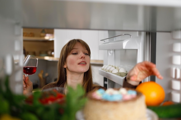 Woman with a glass of red wine in her hand takes an orange from the refrigerator in the kitchen