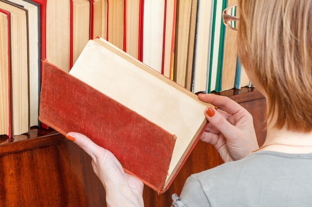 Woman with glasses is holding an old book nex to a bookshelf. Many hardback books on wooden shelf. Library concept