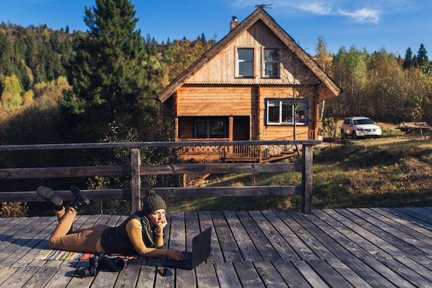 La donna con gli occhiali si trova sul plaid in autunno sulla terrazza in legno e lavora sul computer portatile