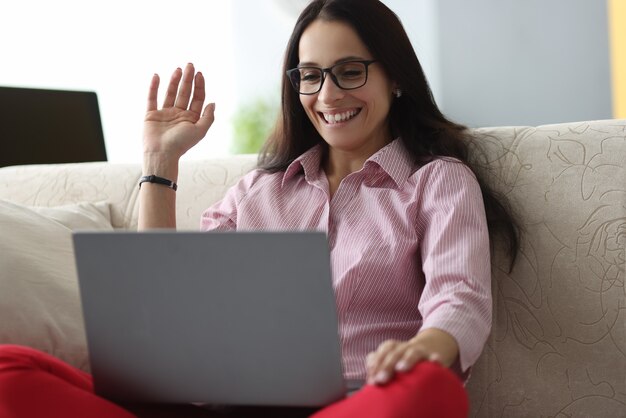 Woman with glasses sits on couch smiles and greets with hand of the interlocutor in the laptop.