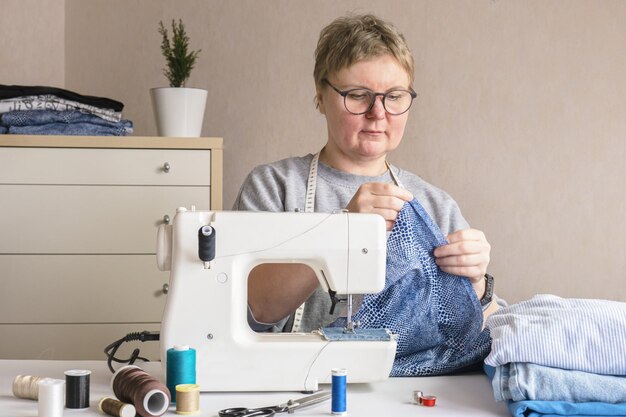 Woman with glasses sits at a table with a sewing machine and sewing supplies A seamstress at her desk sewing clothes Tailor at work