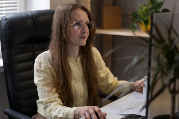 안경을 쓴 여성은 문서와 함께 PC에서 일합니다. 사무실에서 비즈니스 우먼은 작업 문제를 해결합니다.