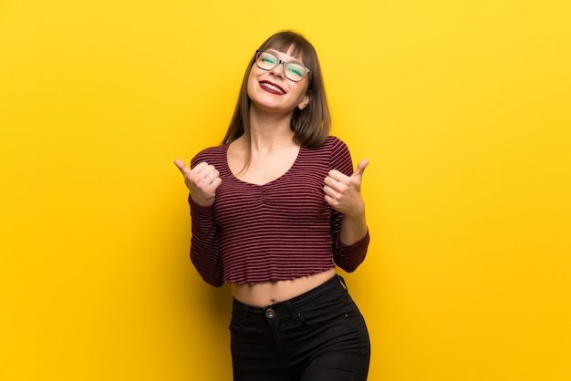 Woman with glasses over yellow wall giving a thumbs up gesture and smiling