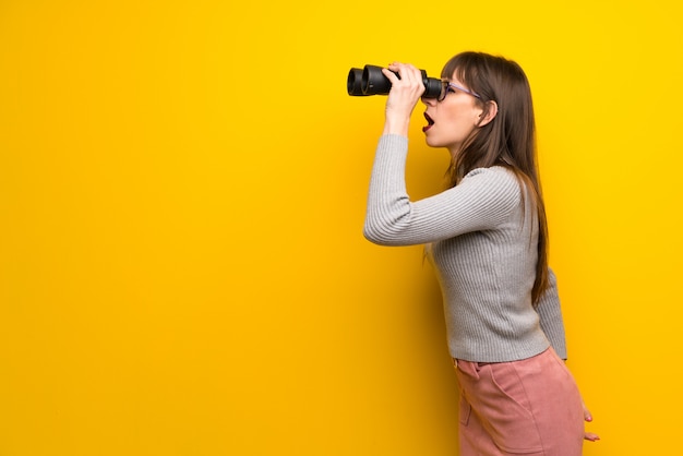 Woman with glasses over yellow wall and looking in the distance with binoculars