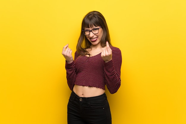 Woman with glasses over yellow wall making money gesture