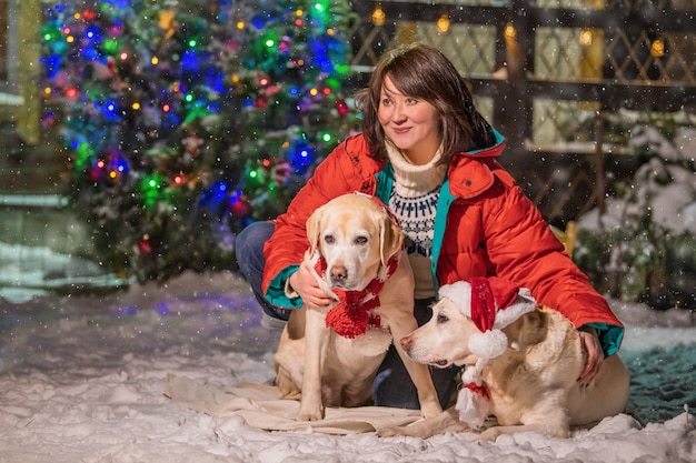 Una donna con un labrador dorato in una sciarpa siede vicino a un albero di natale decorato e a una slitta durante una nevicata in inverno nel cortile di un edificio residenziale.