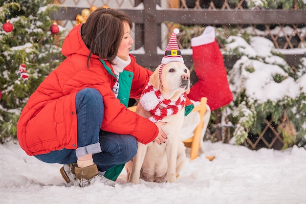 A woman with a golden labrador in a scarf sits near a decorated Christmas tree and sleigh during a snowfall in winter in the courtyard of a residential building.