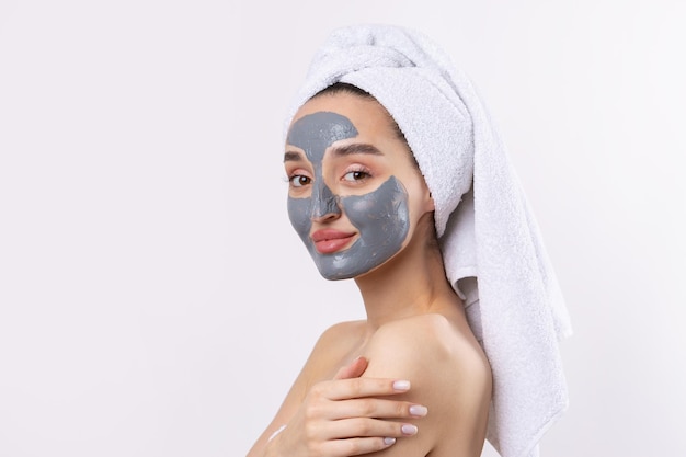 A woman with a gray clay cosmetic mask in a white towel on her head on a white background