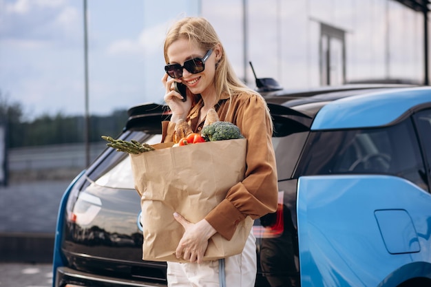 Photo woman with grocery paper bag by her electric car talking on the phone