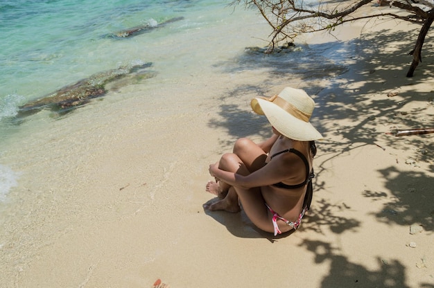 사진 콜롬비아 카르타헤나 섬 해변 앞에 모자와 비키니를 입은 여성이 다리를 꼬고 앉아 있다
