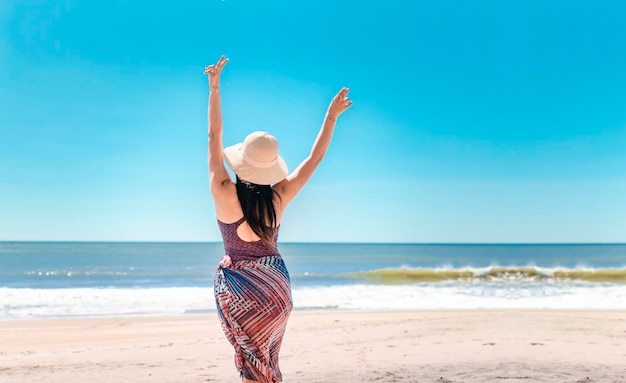 Donna con cappello da dietro sulla spiaggia giovane donna alzando le mani sulla spiaggia