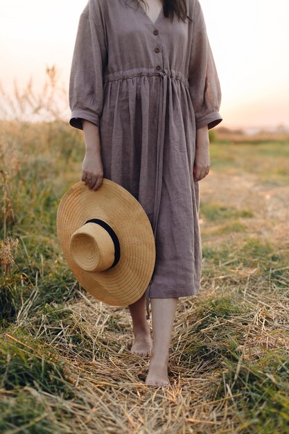 Woman with hat in hand walking barefoot on straw field in sunset light cropped view Rural life