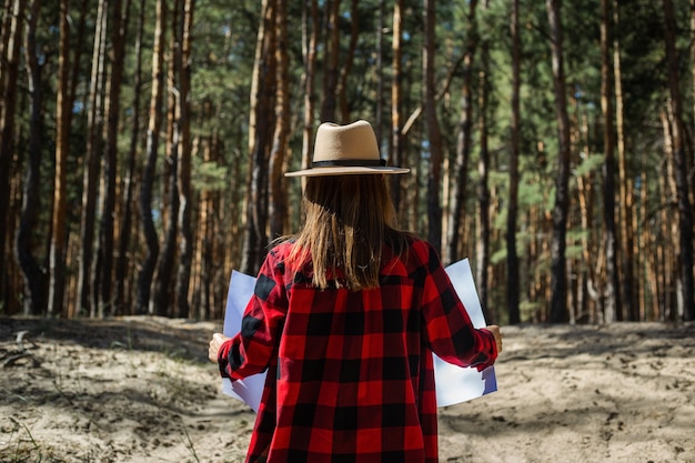 Woman with hat and red plaid shirt holding a map in the forest.