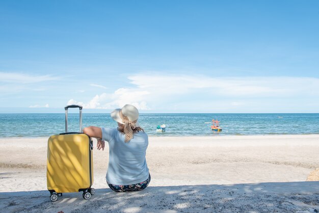 ビーチに旅行のスーツケースで座っている帽子を持つ女性