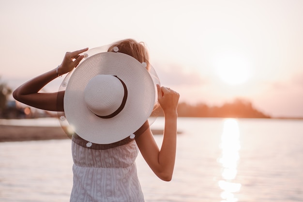 日没後部に帽子をかぶった女性は、夏のサンドレスを着た若いブロンドと装飾された麦わら帽子を見る...