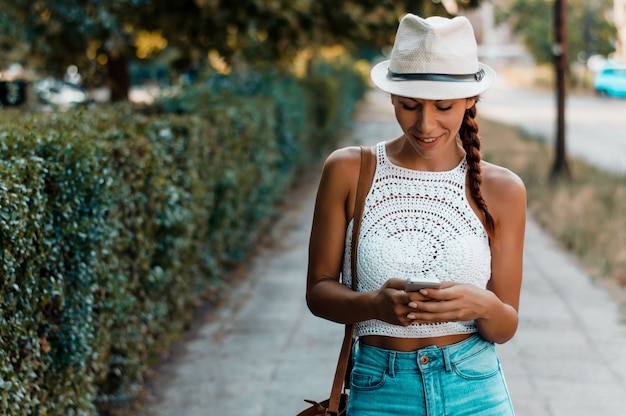 Donna con cappello a piedi e utilizzando uno smart phone in strada in una soleggiata giornata estiva