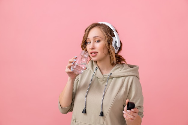 Woman with headphones dressed oversize hoodie drink water glass bottle pink studio background Healthy lifestyle concept