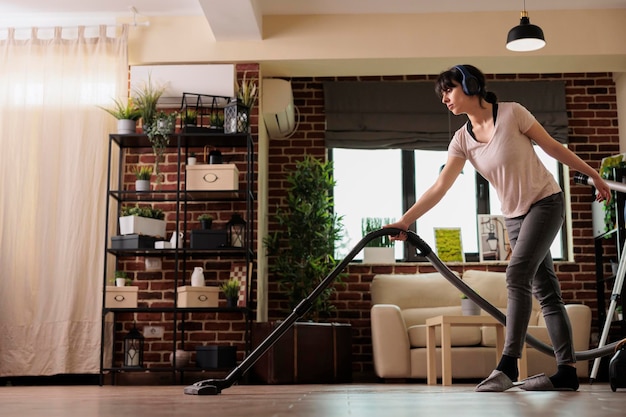 Woman with headphones listening to music doing housework with a modern vacuum cleaner. Housewife enjoying weekly cleaning routine, taking care of household chores in slippers.