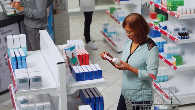 Woman with heart problems checking cardiology pills box, looking for healthcare treatment in pharmacy. Asian person with disease analyzing pharmaceutical products on drugstore shelves. Handheld shot.