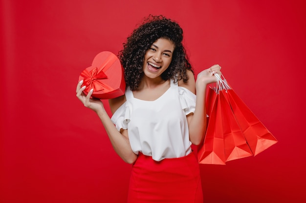 Woman with heart shaped gift box and shopping bags on red wall