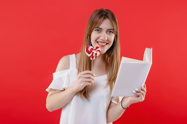 Woman with heart shaped lollypop candy reading book isolated on red wall