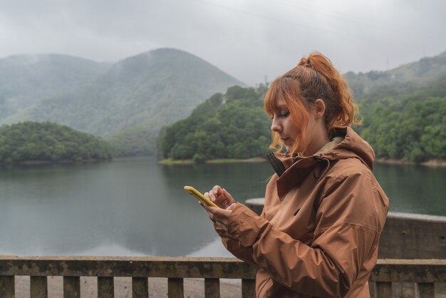 曇りの日に山に囲まれた美しい湖の前でスマートフォンを持った女性