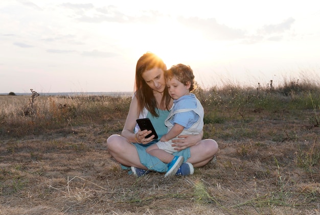 Woman with her threeyearold son in the field at sunset looking at the smartphone connectivity and cover concept