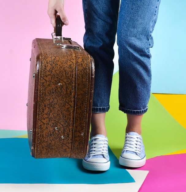 Woman with jeans and sneakers holding in her hand retro suitcase on multicolored paper wall