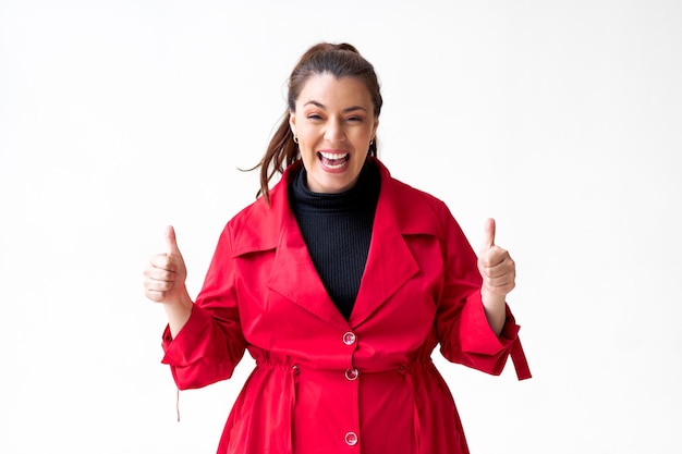 Woman with joyful expression and raised thumbs up wearing red coat on white background