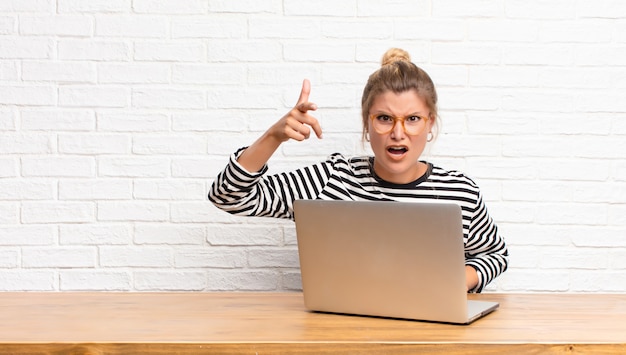 Woman with laptop computer pointing with an angry aggressive expression looking like a furious, crazy boss