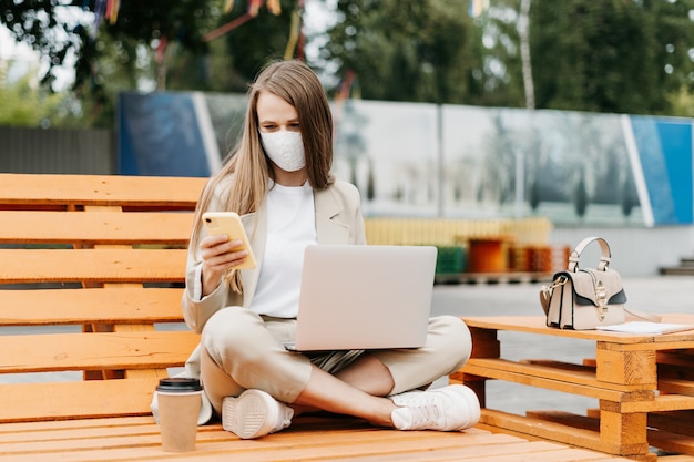Woman with laptop in medical face mask texting over the phone